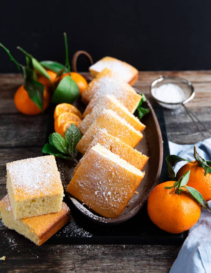 A plate of cut up mandarin orange cake squares dusted with powdered sugar and surrounded by mandarin oranges