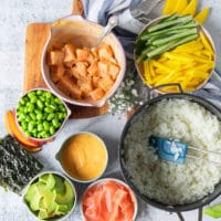 sushi bowl ingredients including a pot with cooked sushi rice, a bowl with salmon and a bowl with spicy mayo, a bowl with edamame , sliced avocado, pickled ginger, some seaweed sheets, a plate with sliced mangoes and cucumbers