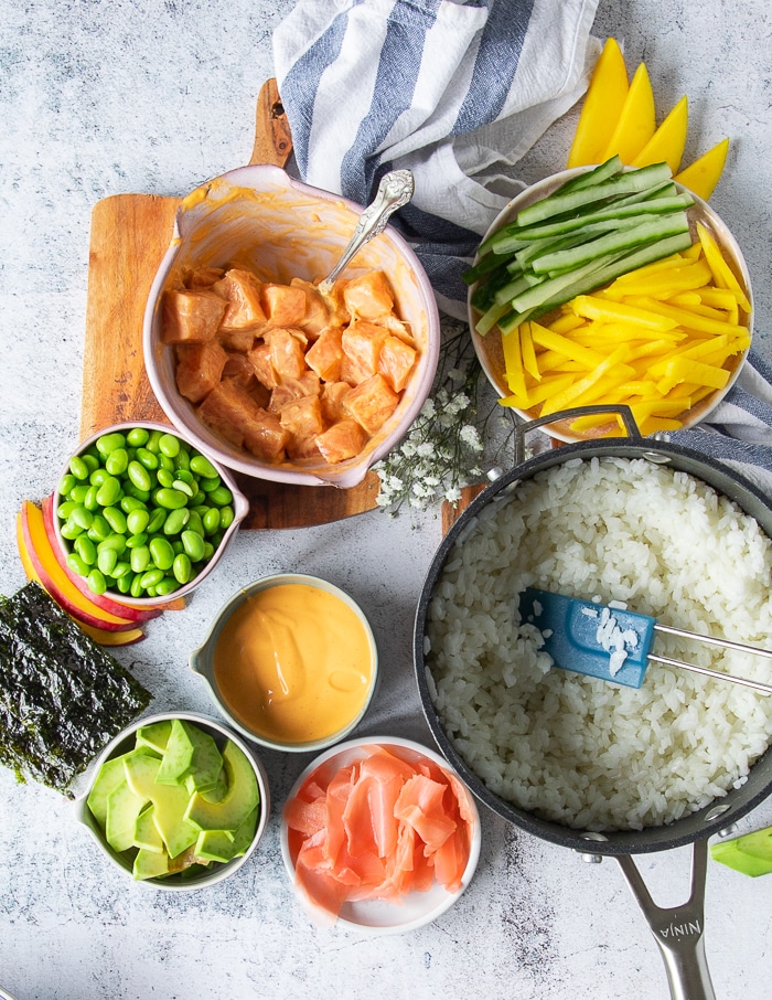 sushi bowl ingredients including a pot with cooked sushi rice, a bowl with salmon and a bowl with spicy mayo, a bowl with edamame , sliced avocado, pickled ginger, some seaweed sheets, a plate with sliced mangoes and cucumbers