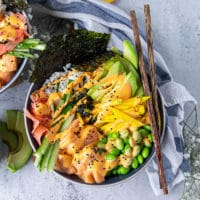 close up of one sushi bowl showing the sushi rice, spicy salmon, edamame , avocado, cucumebrs, mango, ginger, spicy mayo, seaweed and two chopsticks