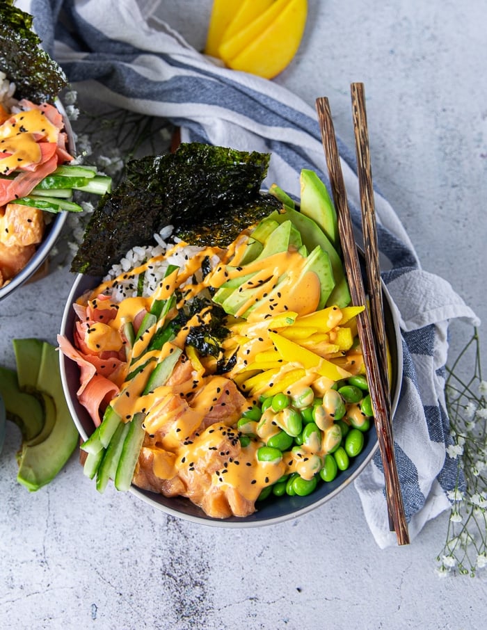 close up of one sushi bowl showing the sushi rice, spicy salmon, edamame , avocado, cucumebrs, mango, ginger, spicy mayo, seaweed and two chopsticks