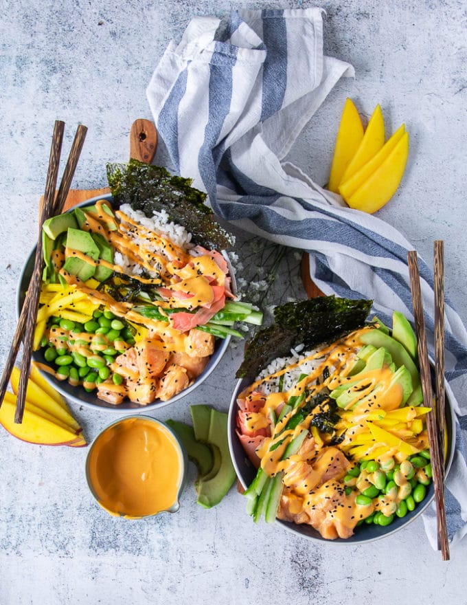 two sushi bowls on a board with wooden sticks. The bowls each have spicy salmon, sushi rice, edamame, avocado, cucumber, mango, pikcled gnger and a drizzle of spicy mayo