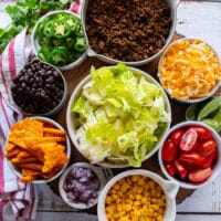 ingredients for taco salad including a bowl of cooked ground beef, some lettuce in a bowl, a bowl of corn, onions, a bowl of tomatoes, some shredded cheese, jalapeños, avocados and chips