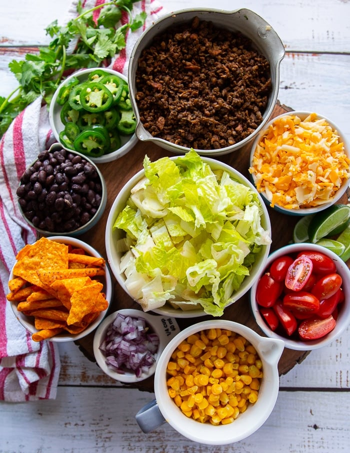 ingredients for taco salad including a bowl of cooked ground beef, some lettuce in a bowl, a bowl of corn, onions, a bowl of tomatoes, some shredded cheese, jalapeños, avocados and chips