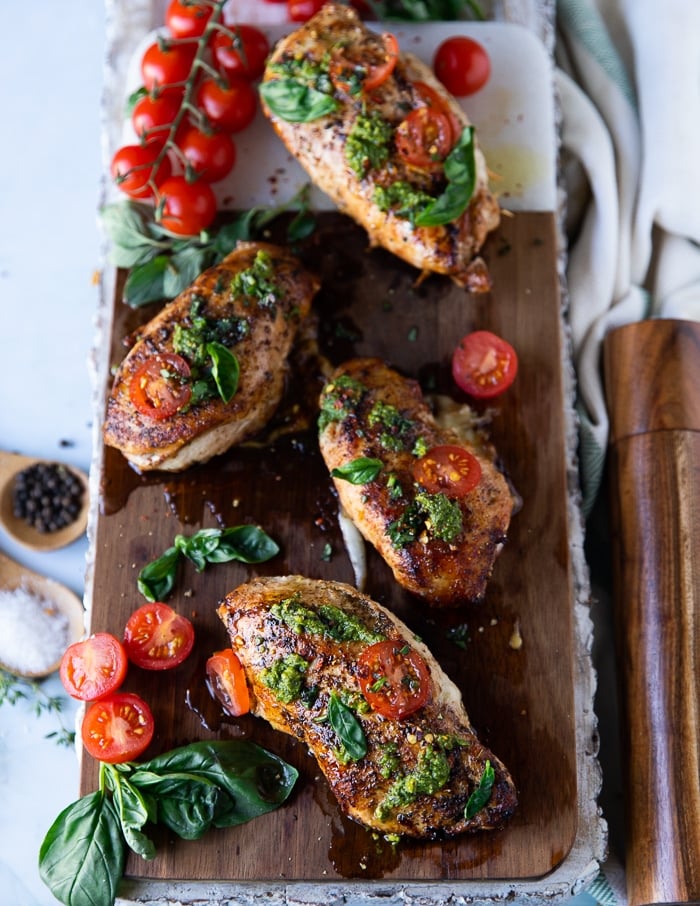 4 pieces of caprese chicken on a wooden board topped with basil pesto and sliced tomatoes, and lots of fresh basil on the side with baby tomatoes