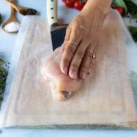 a hand using a sharp knife to cut through the chicken breast and butterfly it