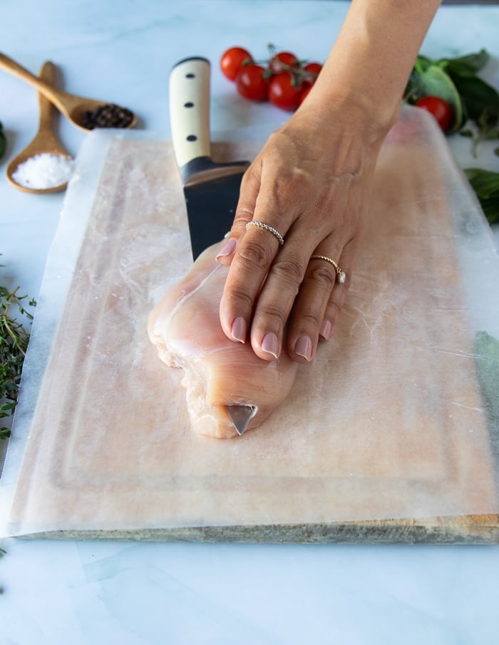 a hand using a sharp knife to cut through the chicken breast and butterfly it 