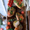 A large wooden board with caprese chicken topped with pesto, tomatoes and stuffed with tomatoes, mozzarella and basil leaves. Once piece of chicken cut in half to show the inside