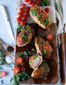 chicken Caprese A large wooden board with caprese chicken topped with pesto, tomatoes and stuffed with tomatoes, mozzarella and basil leaves. Once piece of chicken cut in half to show the inside