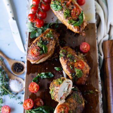 A large wooden board with caprese chicken topped with pesto, tomatoes and stuffed with tomatoes, mozzarella and basil leaves. Once piece of chicken cut in half to show the inside