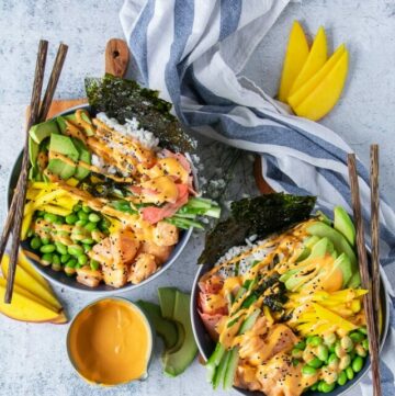 two sushi bowls on a board with wooden sticks. The bowls each have spicy salmon, sushi rice, edamame, avocado, cucumber, mango, pikcled gnger and a drizzle of spicy mayo