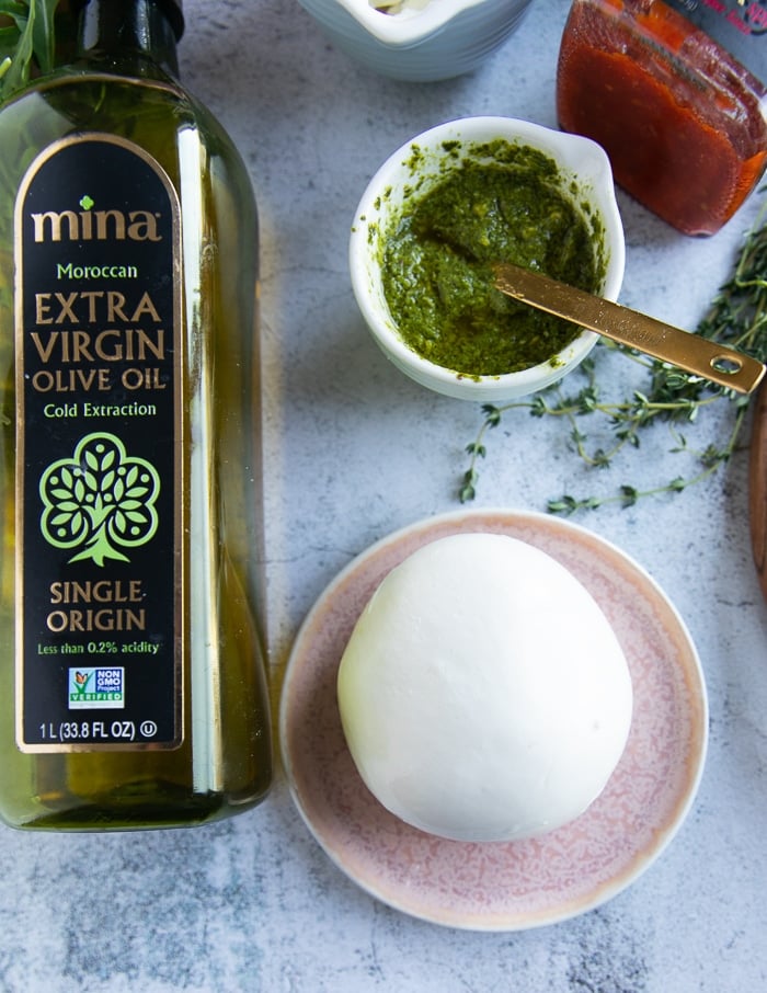 A ball of burrata cheese on a counter next to a bottle of olive oil and some pesto