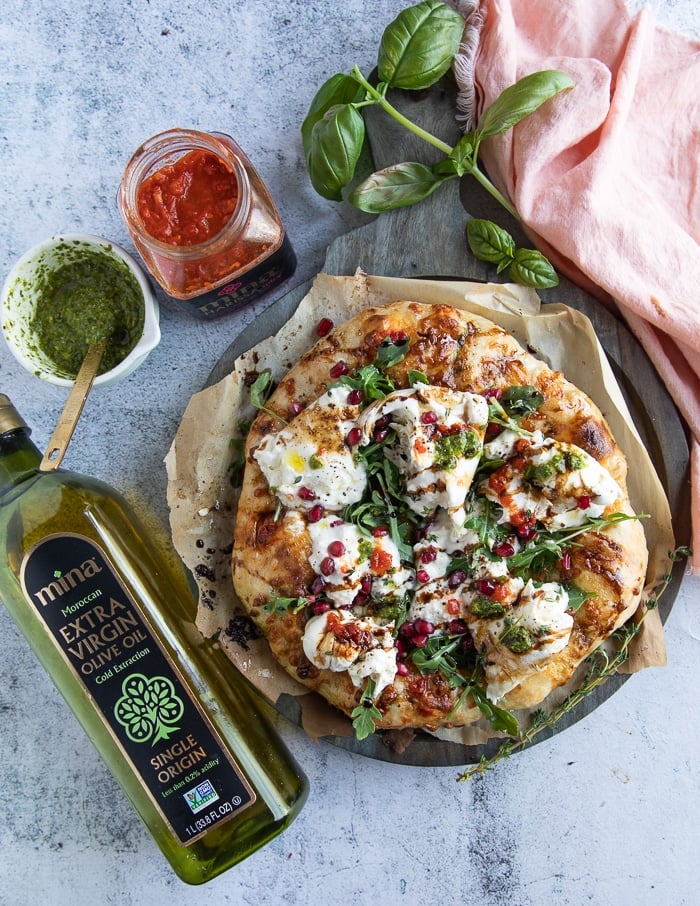 Finished burrata pizza with a jar of olive oil, a jar of harissa, and a small bowl of pesto on the side.