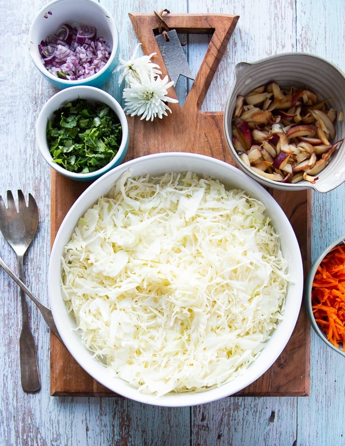 Ingredients for fish taco slaw including cabbage shredded in a bowl, carrots shredded in another bowl, cilantro, diced onions, and shredded apples