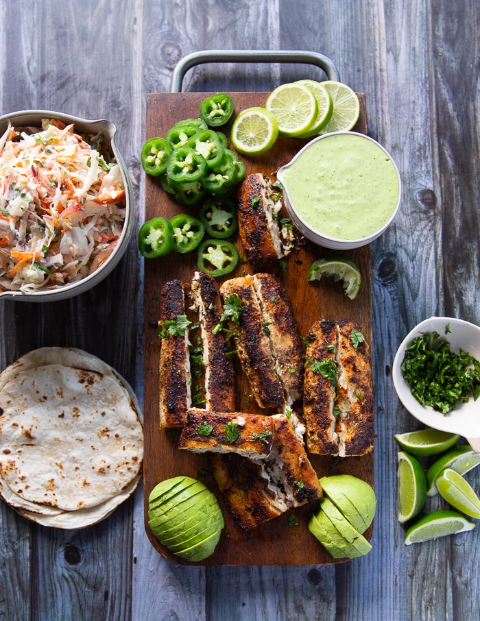 ingredients to assemble the mahi mahi tacos on a board including the cooked mahi mahi fish, fish slaw, poblano sauce, tortilla, jalapenos, avocados, lime wedges and cilantro