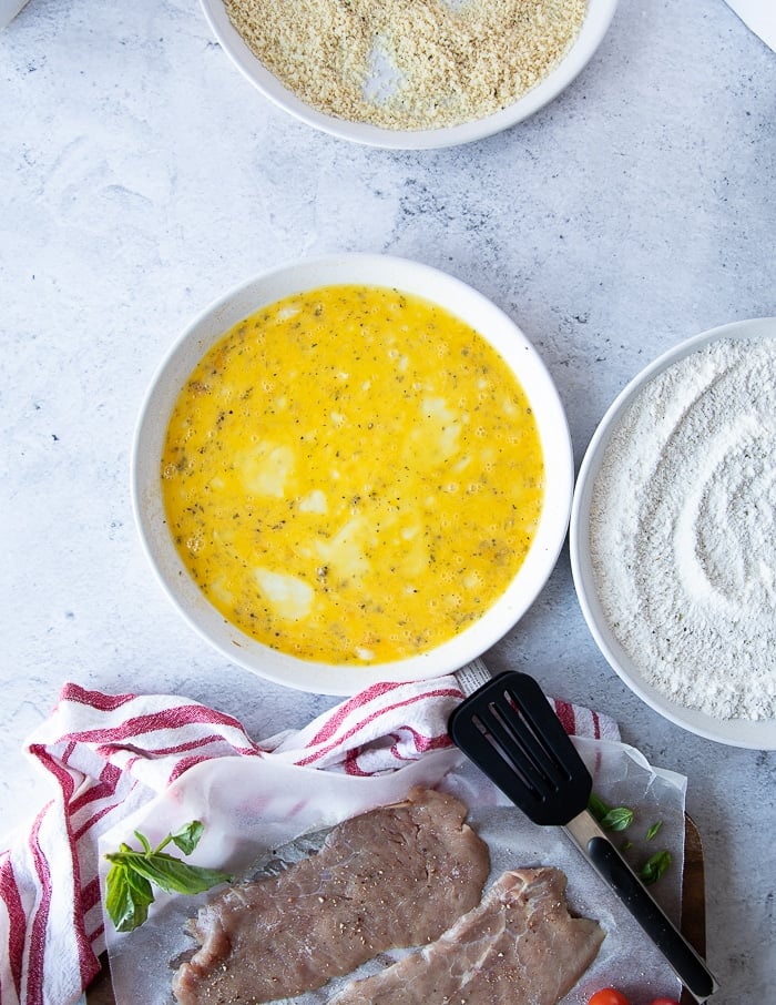 breading station for veal parmesan recipe including a plate of seasoned flour, plate of seasoned eggs, plate of seasoned breadcrumbs