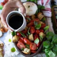 a hand holding a small pouring jug of balsamic syrup mixed with olive oil and about to pour it over the burrata caprese