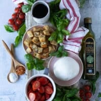 ingredients for burrata caprese including a plate of burrata cheese, a plate of garlic croutons, a bowl of cherry tomatoes cut in half, lots of fresh basil leaves, a bottle of olive oil