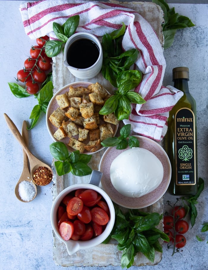 ingredients for burrata caprese including a plate of burrata cheese, a plate of garlic croutons, a bowl of cherry tomatoes cut in half, lots of fresh basil leaves, a bottle of olive oil