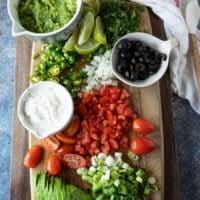 a board with a variety of toppings for the carne asada fries recipe including tomatoes, avocado, scallions, sour cream, black olives, jalapenos, cilantro and more