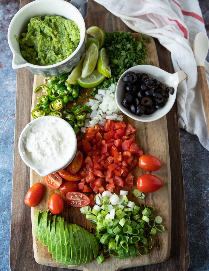 a board with a variety of toppings for the carne asada fries recipe including tomatoes, avocado, scallions, sour cream, black olives, jalapenos, cilantro and more