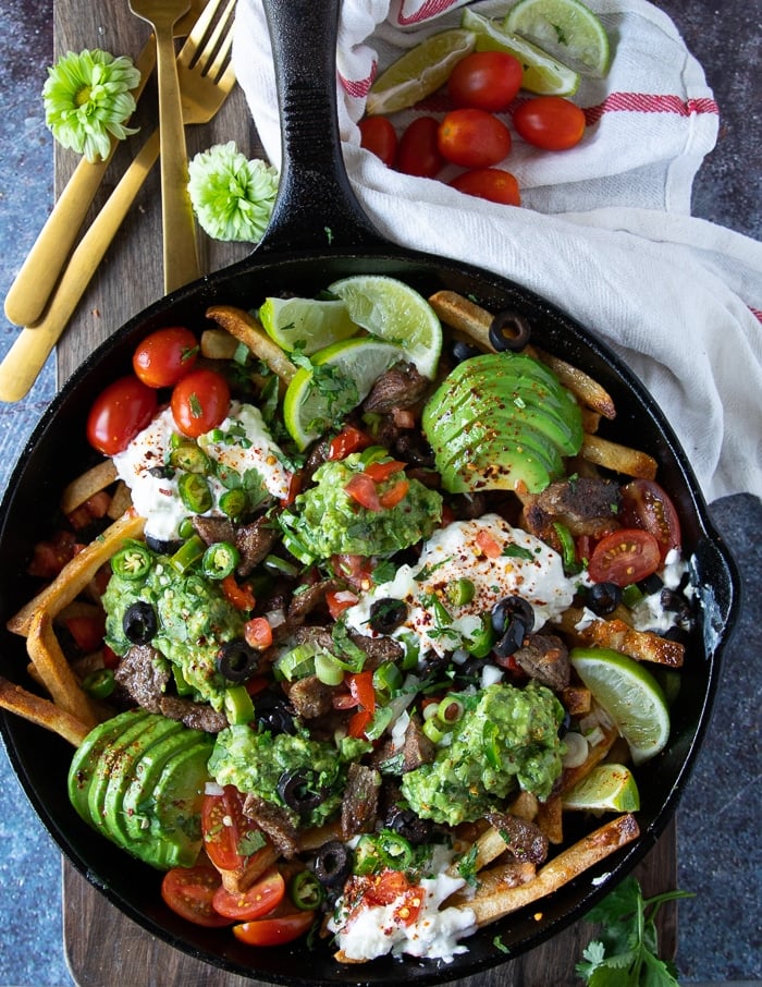 carne asada fries in a skillet topped with meat, cheese, avocado slices, sour cream, tomatoes, olives and lime wedges