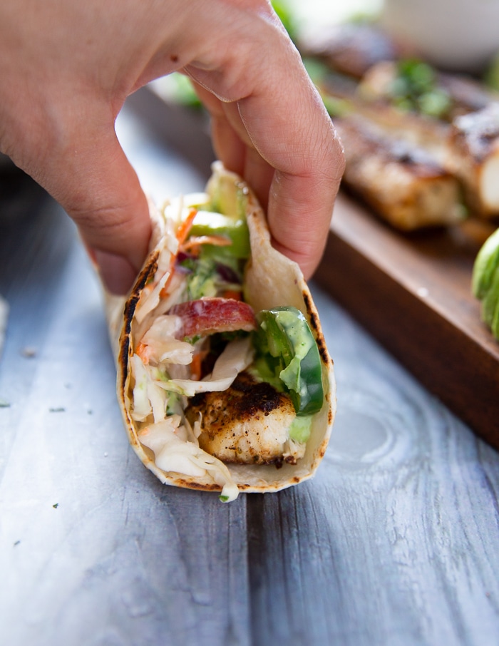 A hand holding the finished mahi mahi tacos sideways showing the mahi mahi fish, fish slaw, avocado and poblano sauce