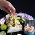 A hand holding a piece of mahi mahi tacos on a board near a bowl of fish slaw