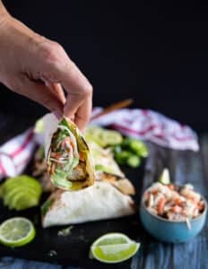 A hand holding a piece of mahi mahi tacos on a board near a bowl of fish slaw