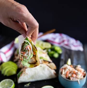 A hand holding a piece of mahi mahi tacos on a board near a bowl of fish slaw