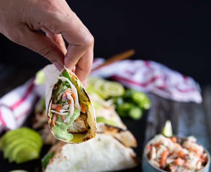 A hand holding a piece of mahi mahi tacos on a board near a bowl of fish slaw