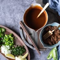 Ingredients for Birria ramen including a bowl of birria consome, birria meat, cooked noodles, toppings such as cilantro, jalapenos, diced onions and lime wedges