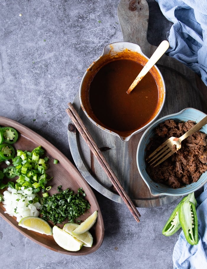 Ingredients for Birria ramen including a bowl of birria consome, birria meat, cooked noodles, toppings such as cilantro, jalapenos, diced onions and lime wedges