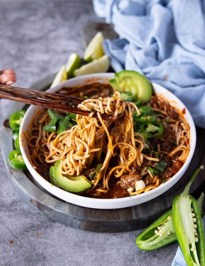 two chopsticks stretching out the birria ramen showing the rich birria stew and birria meat drenching the noodles with al the toppings