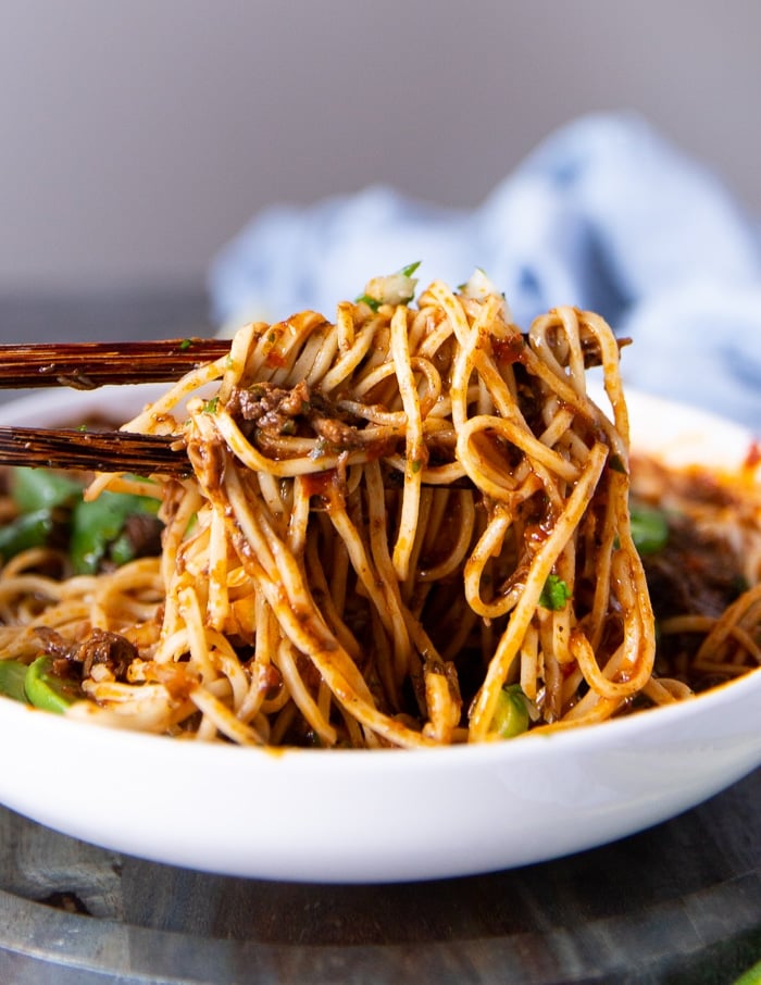 two chopsticks lifting up the noodles close up showing the birria ramen coated in so much sauce and the meat shreds with the toppings