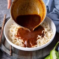 A hand pouring the Birria consommé over the cooked noodles