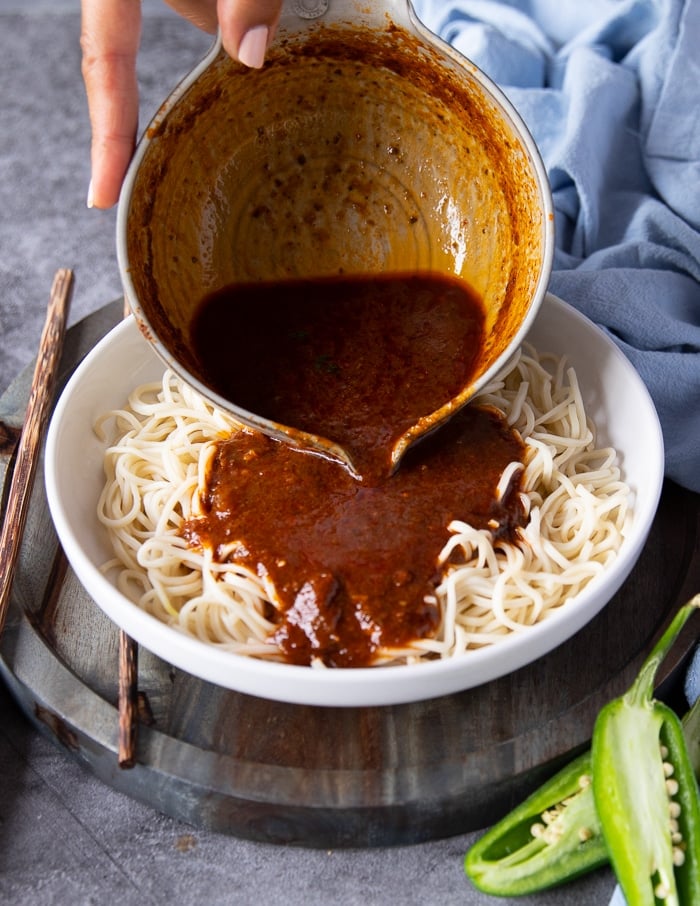 A hand pouring the Birria consommé over the cooked noodles