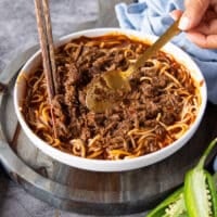 A hand adding lots of shredded birria meat over the noodles