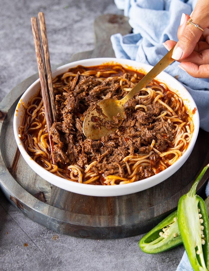 A hand adding lots of shredded birria meat over the noodles