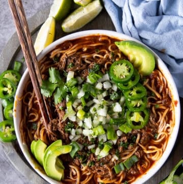 Top view of a bowl of birria ramen with two chopsticks and topped with avocados, jalapenos, onions and cilantro