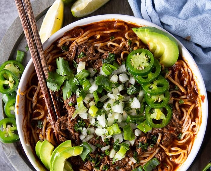 Top view of a bowl of birria ramen with two chopsticks and topped with avocados, jalapenos, onions and cilantro