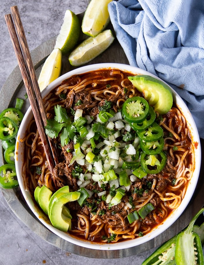 Top view of a bowl of birria ramen with two chopsticks and topped with avocados, jalapenos, onions and cilantro