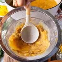 A hand using a wooden spoon to mash the apple cider mixture firmly over the strainer so that the homemade apple cider is pulp free