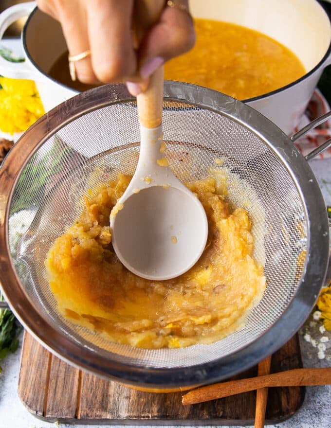 A hand using a wooden spoon to mash the apple cider mixture firmly over the strainer so that the homemade apple cider is pulp free