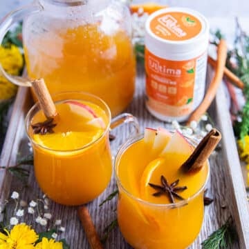 A tray with two cups of homemade apple cider and a large glass jar of apple cider to serve extras, surrounded by flowers, and garnished with cinnamon sticks, orange slices, apple slices