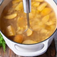 A hand holding a potato masher and mashing down the apples and oranges in the pot to make homemade apple cider