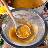 a hand holding a ladle of homemade apple cider and placing it over a fine mesh strainer