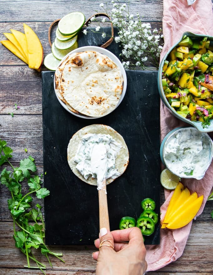 Assembling the tacos recipe: hand spreading some herb sour cream over a tortilla