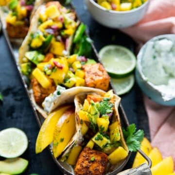 close up of salmon tacos on a plate with some lime slices around, some sour cream and avocado salsa
