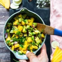 A hand holding a bowl of avocado salsa being prepared while the salmon is cooking